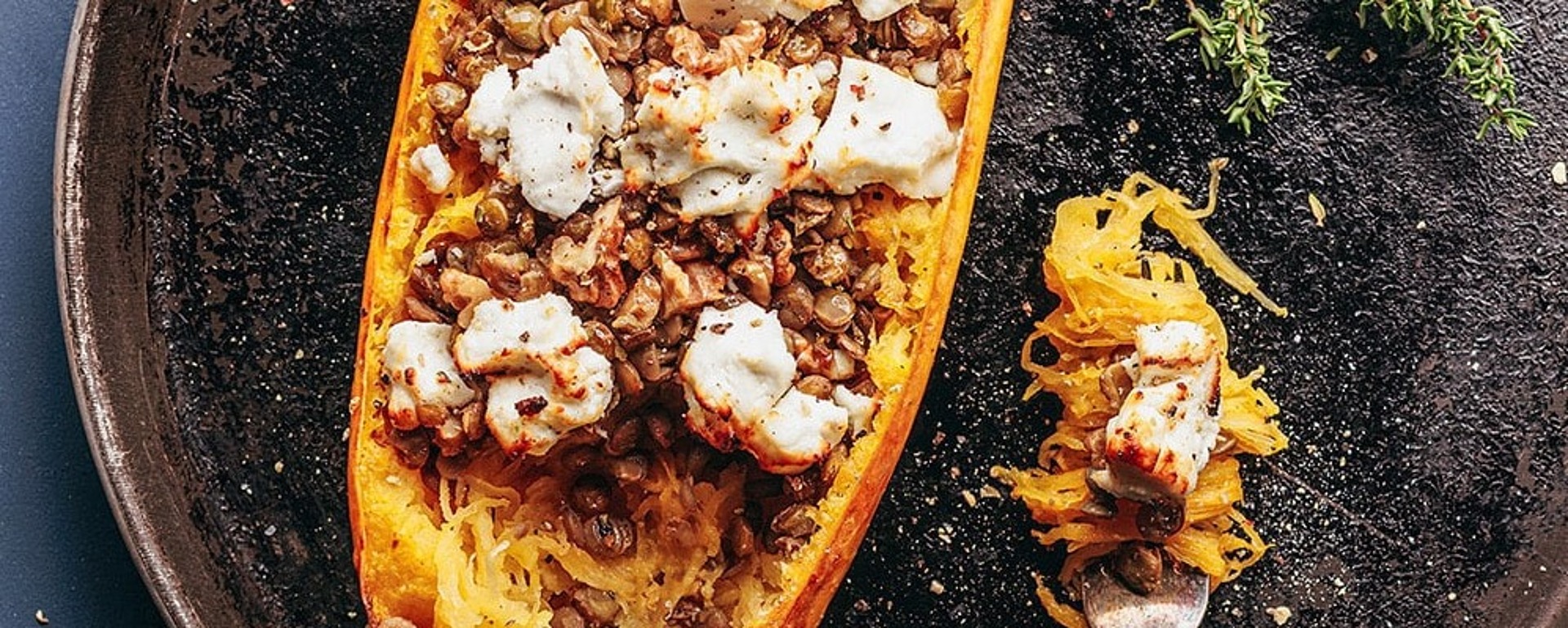 Ein gebratener halber Spaghettikürbis, gefüllt mit Linsen und überbacken mit geschmolzenem Käse, liegt auf einem runden Backblech, daneben frischer Thymian. Auf dem Tablett liegt eine Gabel, auf der ein Bissen des Gerichts liegt.