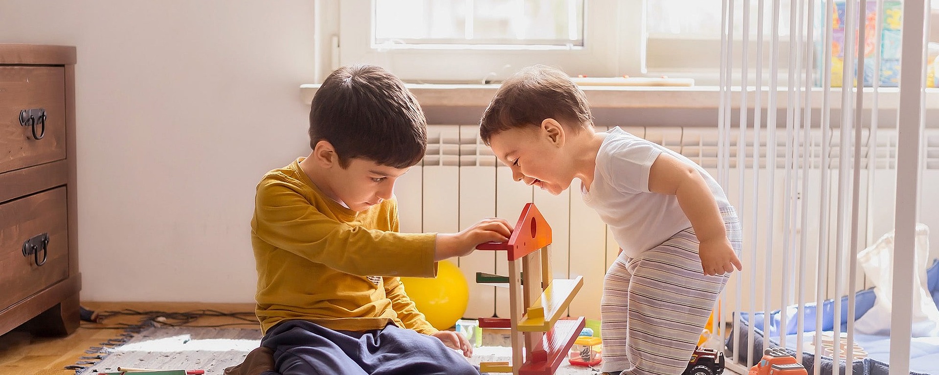Zwei kleine Kinder spielen zusammen auf einem Teppich in einem sonnenbeschienenen Raum mit einem bunten Holzspielzeug. Ein Kind sitzt konzentriert, während das andere steht und sich neugierig anlehnt. Spielzeug und Bücher sind um sie herum verstreut.