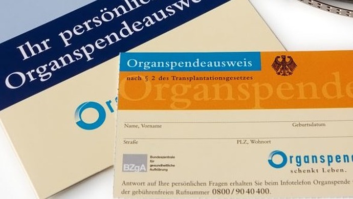 Ein deutscher Organspendeausweis und eine Informationsbroschüre sind in Nahaufnahme auf einer weißen Fläche abgebildet. Die Dokumente sind mit blauen und orangefarbenen Akzenten versehen.