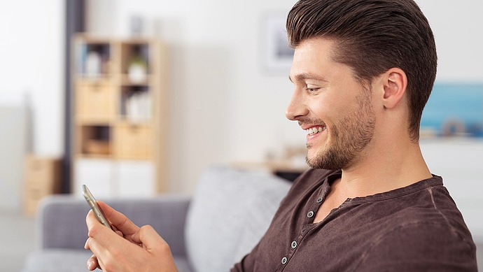 Ein junger Mann mit kurzen braunen Haaren und einem gestutzten Bart sitzt lächelnd auf einer Couch und schaut auf sein Smartphone. Der Hintergrund zeigt einen verschwommenen Raum mit Regalen und einem Gemälde.