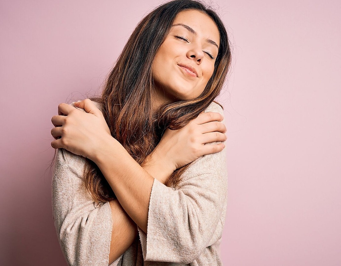 Eine junge Frau mit langen braunen Haaren umarmt sich selbst und lächelt mit geschlossenen Augen, während sie vor einem rosa Hintergrund steht. Sie sieht zufrieden und entspannt aus und trägt einen hellen Pullover.
