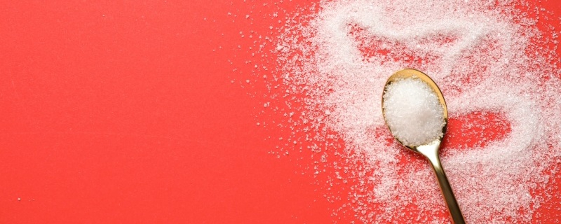 Ein mit Kristallzucker gefüllter Löffel liegt auf einer leuchtend roten Oberfläche. Etwas Zucker ist um den Löffel herum verstreut und bildet eine grobe Herzform. Der Kontrast zwischen dem weißen Zucker und dem roten Hintergrund ist auffällig und erzeugt ein optisch ansprechendes Bild.