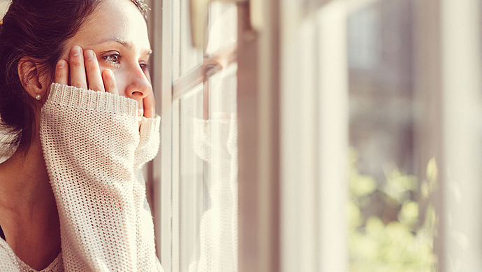 Eine Frau blickt nachdenklich aus dem Fenster. Ihre Hände ruhen sanft auf ihren Wangen. Sie trägt einen kuscheligen, hellen Pullover, und natürliches Licht erhellt ihr Gesicht sanft. Der Hintergrund ist unscharf und suggeriert eine ruhige Szene im Freien.
