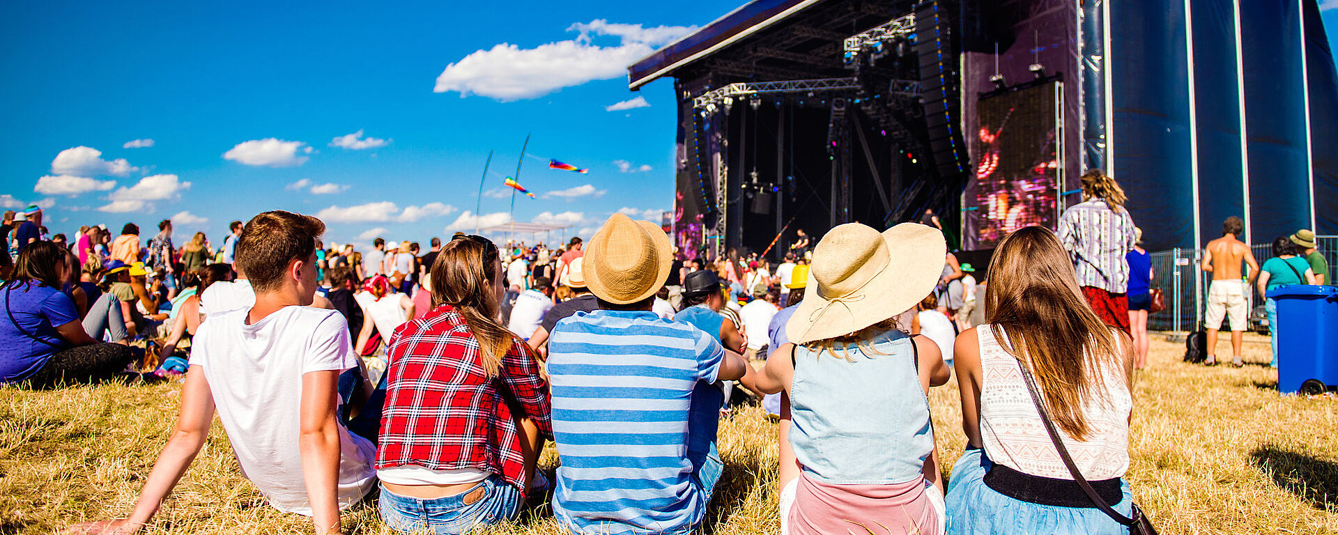 Eine Gruppe von Menschen sitzt auf einer Wiese vor einer Freilichtbühne bei einem Musikfestival. Im Hintergrund sind eine Menschenmenge, bunte Fahnen und ein strahlend blauer Himmel zu sehen. Einige tragen Hüte und sommerliche Kleidung.