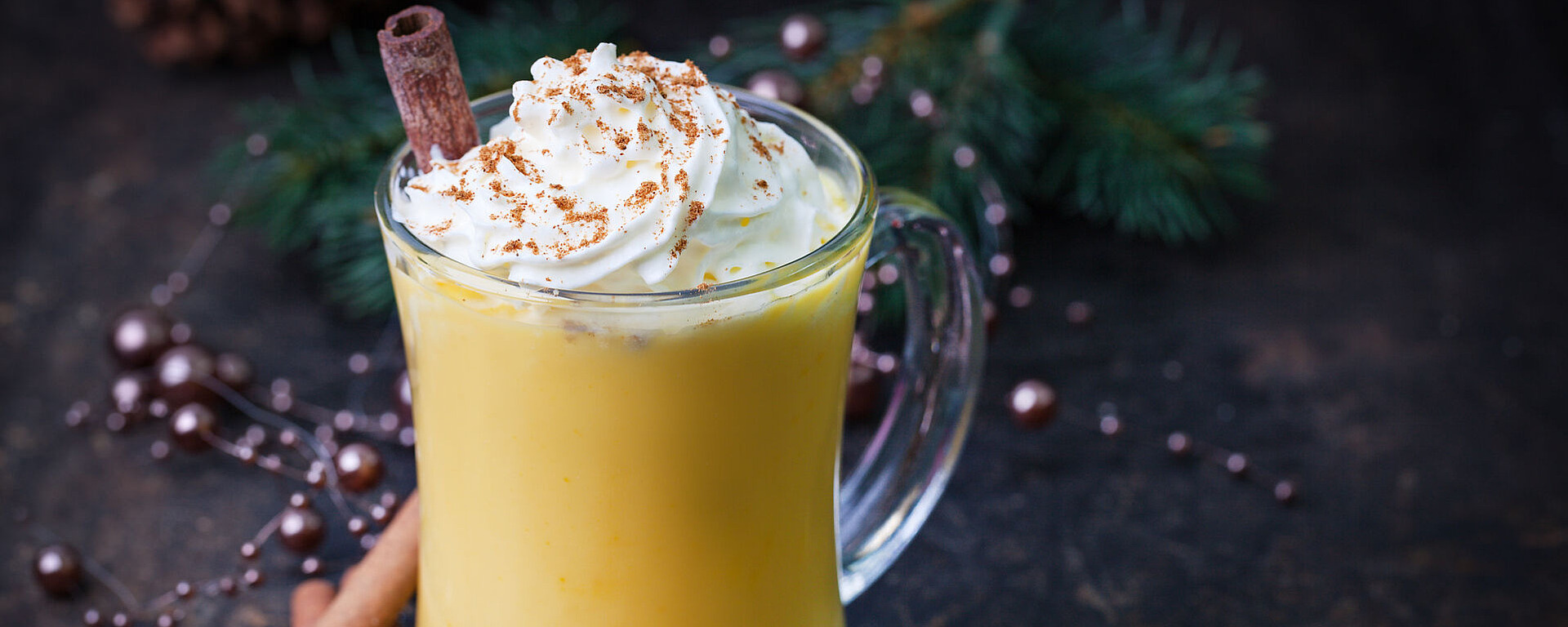 Ein Glasbecher mit Eierlikör, gekrönt mit Schlagsahne und einer Prise Gewürzen, garniert mit einer Zimtstange. Ganze Zimtstangen und Sternanis stehen neben dem Becher, mit festlichem Grün im Hintergrund.