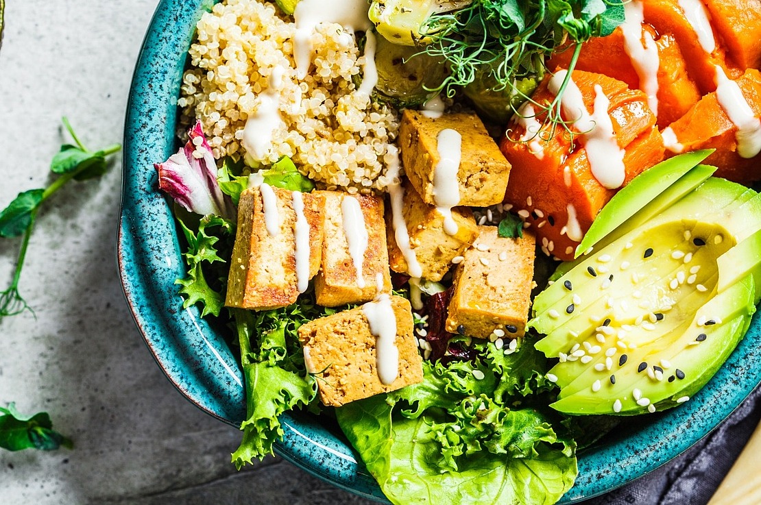Eine farbenfrohe Schüssel mit Quinoa, gegrillten Tofuwürfeln, Blattgemüse, in Scheiben geschnittener Avocado mit Sesam, gerösteten Zucchini und Orangengemüse, garniert mit einem Spritzer cremigem Dressing.