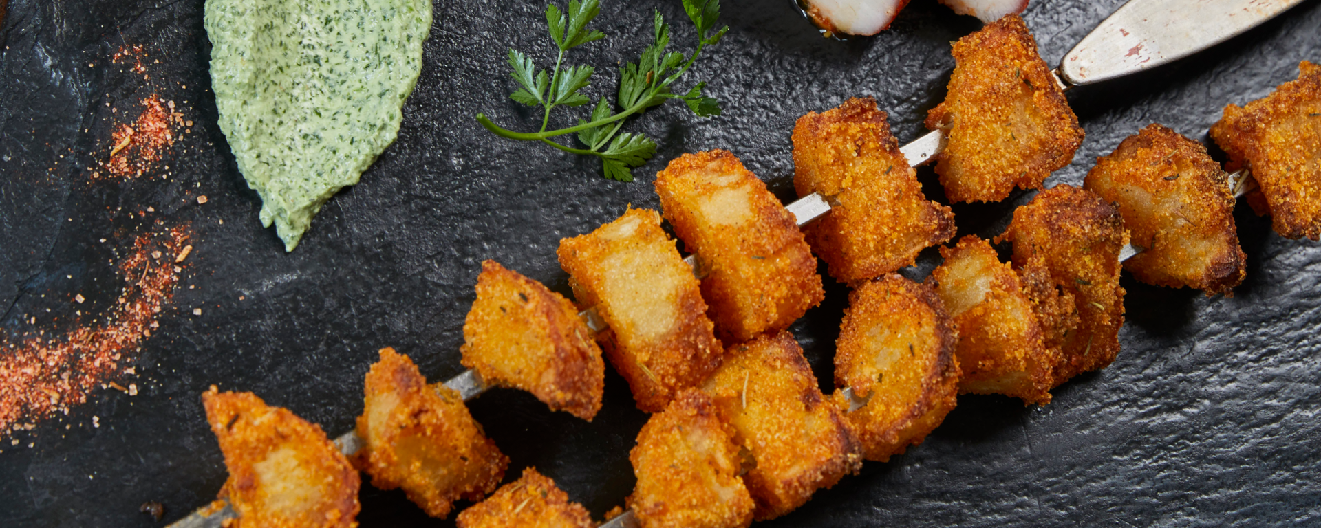 Ein Spieß mit goldenen, knusprig gebratenen Würfeln liegt auf einer Schieferplatte neben gebratenen Fleischscheiben, einem Klecks grüner Soße, frischer Kräutergarnitur und einer Prise Gewürzen.