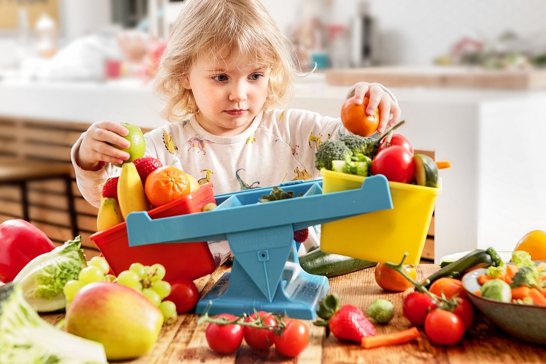 Kind spielt mit einer Waage und balanciert Obst und Gemüse darauf