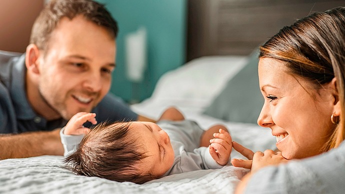 Ein lächelndes Paar liegt auf einem Bett und betrachtet liebevoll sein neugeborenes Baby, das zwischen ihnen liegt und seine kleinen Hände nach oben streckt. Die Szene ist warm und unterstreicht die familiäre Verbundenheit.