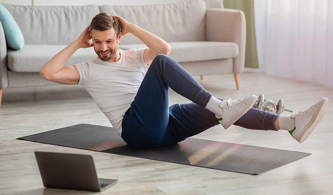 Ein Mann trainiert zu Hause auf einer Yogamatte und macht ein Workout auf seinem Laptop. Er macht einen Bicycle Crunch und trägt ein weißes T-Shirt, blaue Hosen und weiße Turnschuhe. In der Nähe liegen Hanteln auf dem Boden und im Hintergrund steht ein Sofa.
