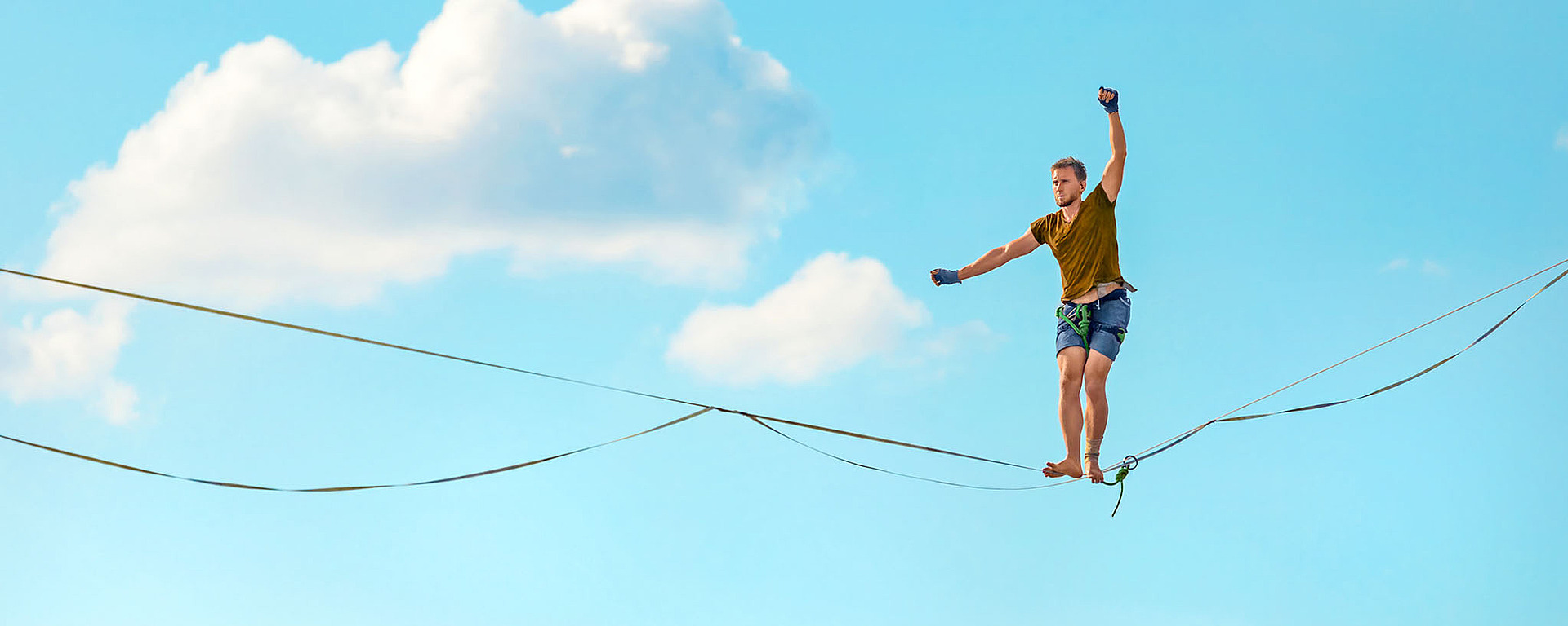 Eine Person balanciert barfuß auf einer Slackline hoch über dem Boden vor einem strahlend blauen Himmel mit vereinzelten Wolken und hat die Arme zur Stabilisierung ausgestreckt.