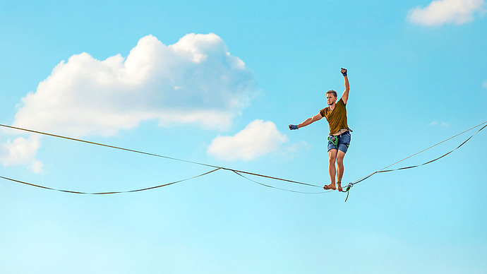 Eine Person balanciert barfuß auf einer Slackline hoch über dem Boden vor einem strahlend blauen Himmel mit vereinzelten Wolken und hat die Arme zur Stabilisierung ausgestreckt.