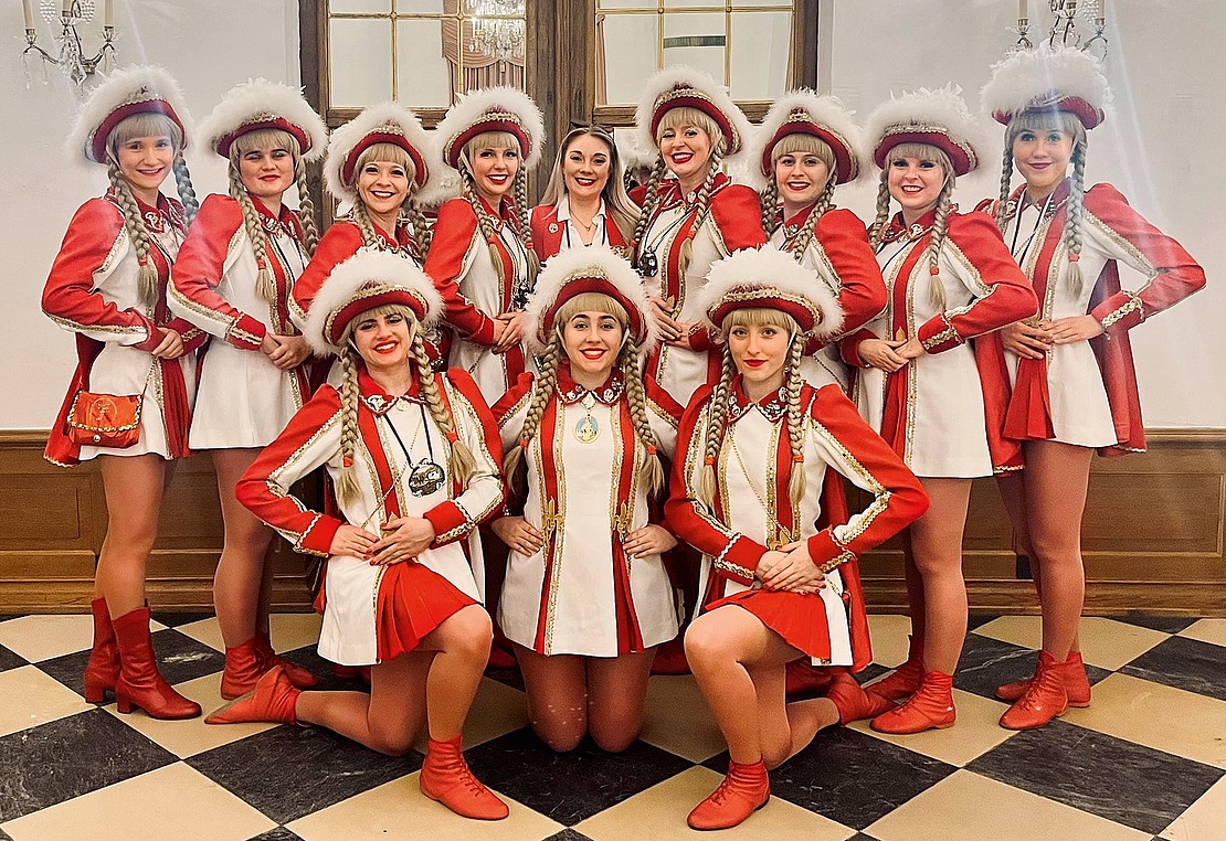Zwölf Frauen in passenden rot-weißen Uniformen mit Federhüten posieren in zwei Reihen lächelnd auf einem karierten Boden vor einem großen Fenster und zwei Wandleuchtern.