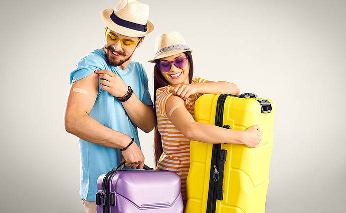 Ein lächelnder Mann und eine lächelnde Frau in Sommerkleidung und Hüten zeigen Verbände an ihren Armen und halten lila und gelbe Koffer in den Händen, was darauf hindeutet, dass sie vor der Reise gerade ihre Impfungen erhalten haben.