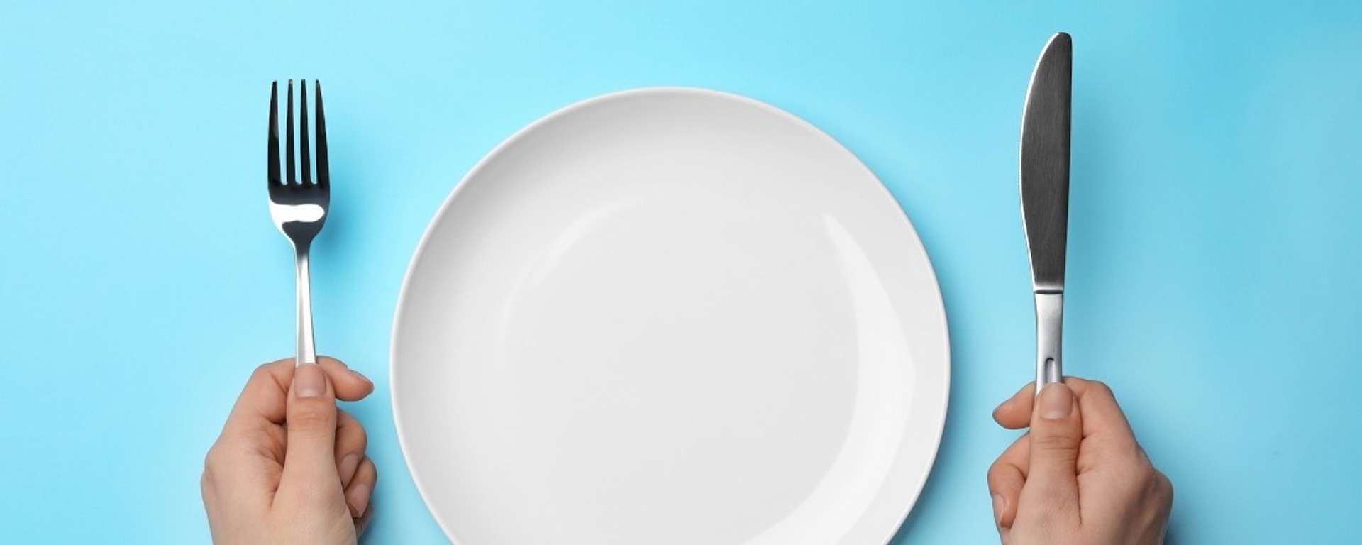 Eine Person hält in der linken Hand eine Gabel und in der rechten Hand ein Messer, die auf beiden Seiten eines leeren weißen Tellers vor einem blauen Hintergrund liegen.