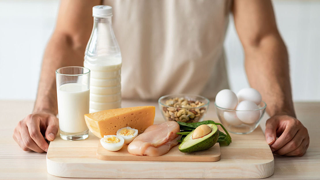 Eine Person steht hinter einem Schneidebrett, auf dem Eier, Avocado, Käse, Hähnchenbrust, Nüsse sowie Gläser und eine Flasche Milch ordentlich angeordnet sind.