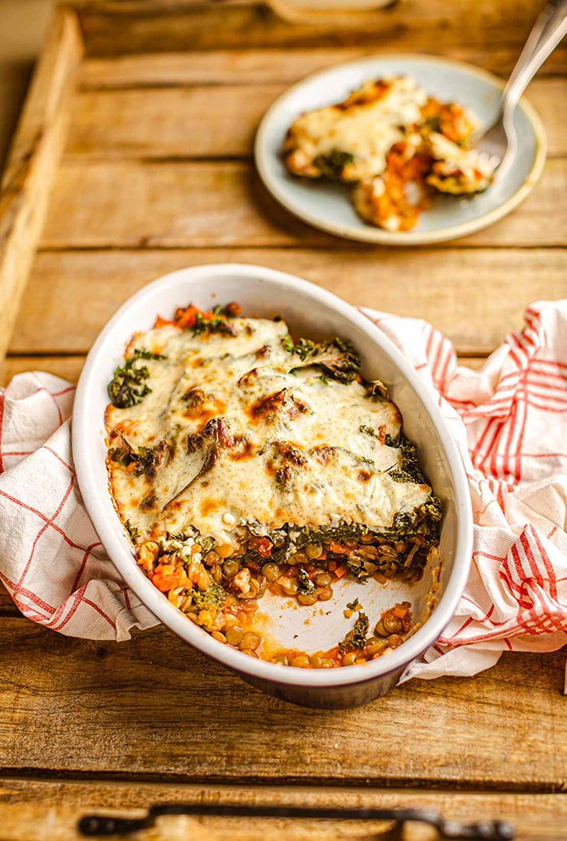 Ein Holztisch mit einer Auflaufform, die mit einem überbackenen Gemüsegericht mit Käse, Grünzeug und Karotten gefüllt ist. Unter der Form liegt ein rot-weißes Geschirrtuch und im Hintergrund ein kleiner Teller mit einer Portion und einer Gabel.