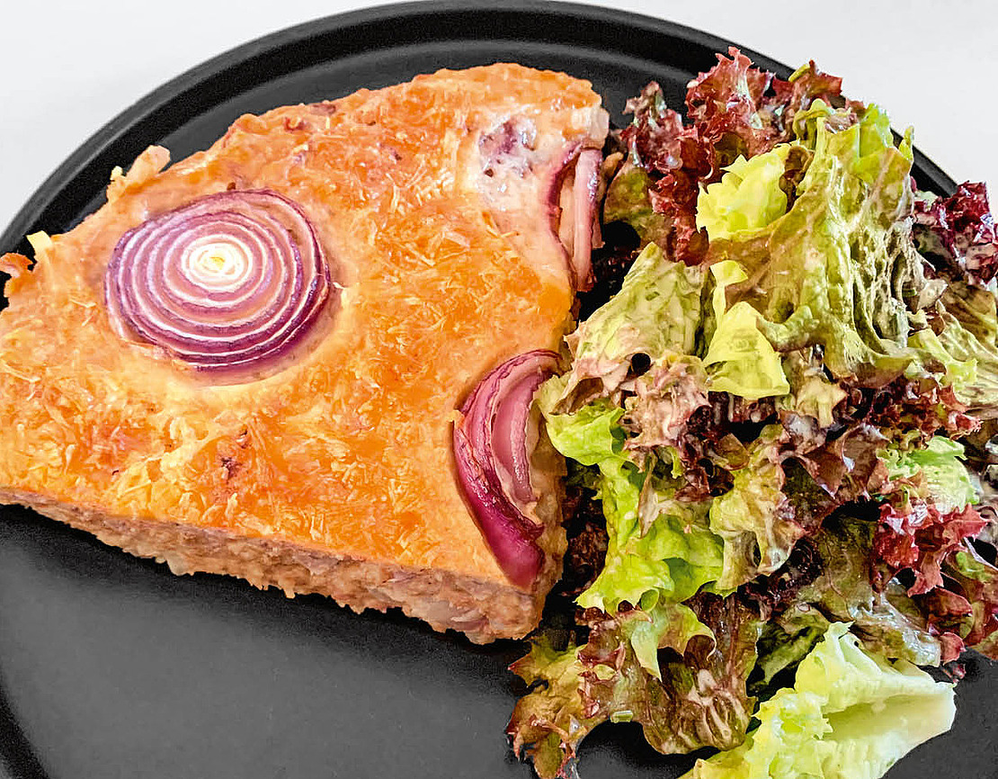 Ein Stück herzhafte Pastete mit sichtbaren roten Zwiebelringen obenauf, serviert auf einem schwarzen Teller zusammen mit einer Portion gemischtem Blattsalat.