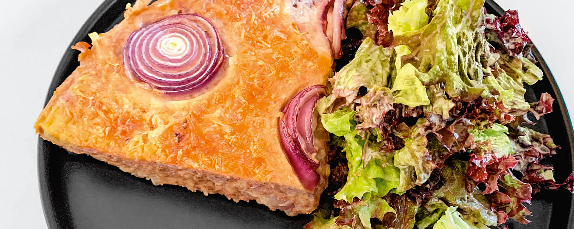 Ein Stück herzhafte Pastete mit sichtbaren roten Zwiebelringen obenauf, serviert auf einem schwarzen Teller zusammen mit einer Portion gemischtem Blattsalat.