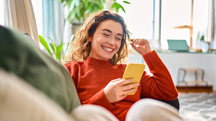 Eine junge Frau mit lockigem Haar, die einen roten Pullover trägt, lächelt, während sie auf ihr gelbes Smartphone schaut. Sie sitzt gemütlich in einem Raum mit Pflanzen und Sonnenlicht im Hintergrund.