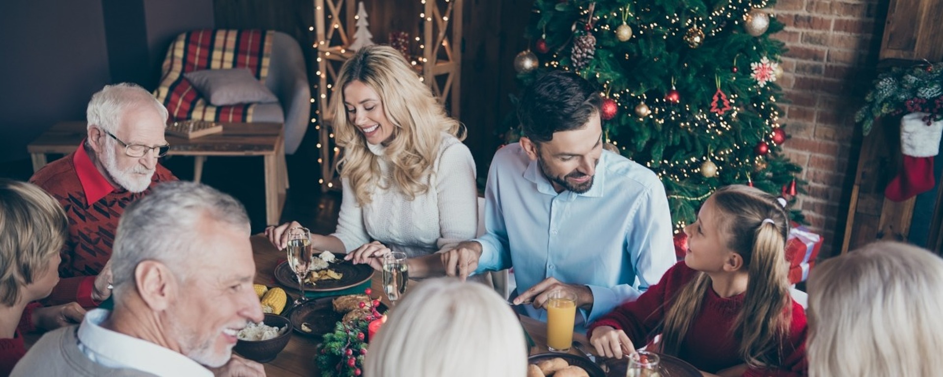 Eine achtköpfige Familie versammelt sich um einen Esstisch, der mit festlichen Weihnachtselementen dekoriert ist. Im Hintergrund steht ein Weihnachtsbaum. Alle lächeln und genießen gemeinsam das Essen.