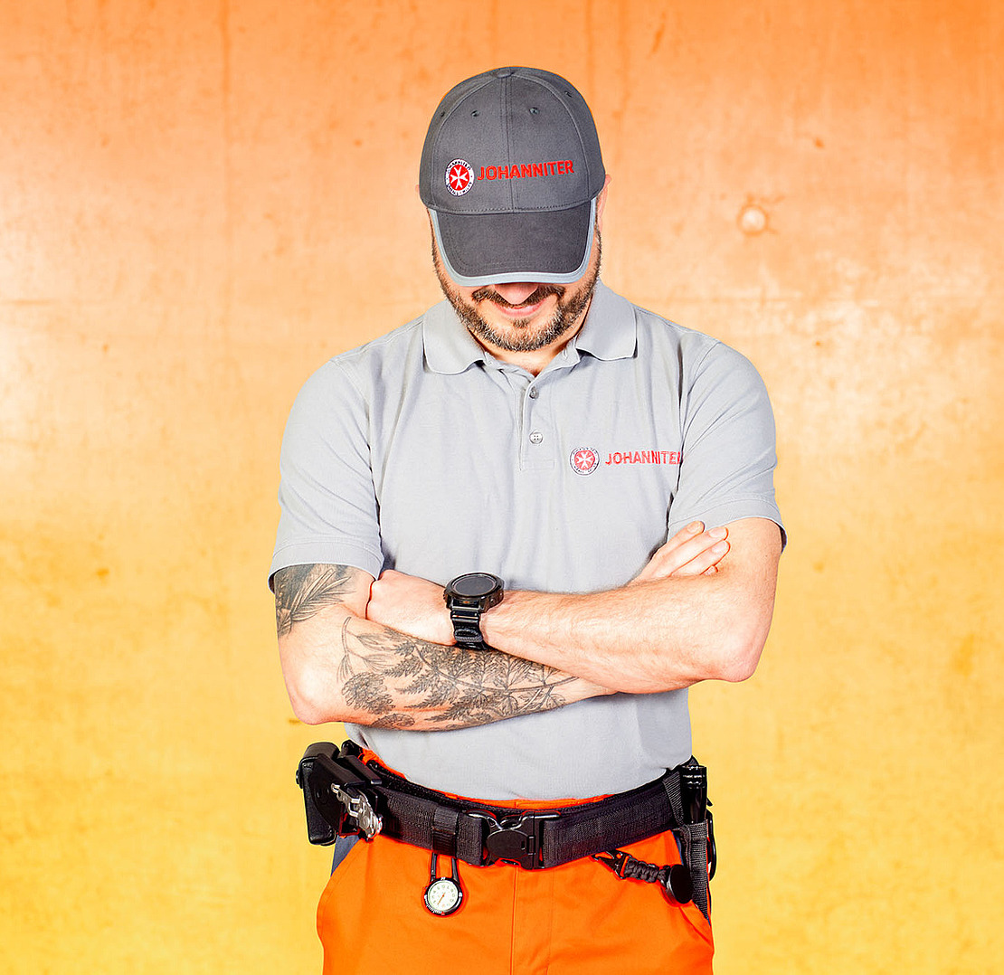 Ein Mann mit grauer Johannitermütze und Poloshirt steht mit verschränkten Armen vor orangefarbenem Hintergrund und zeigt eine Tätowierung auf seinem rechten Unterarm. Er trägt einen Werkzeuggürtel und blickt nach unten.