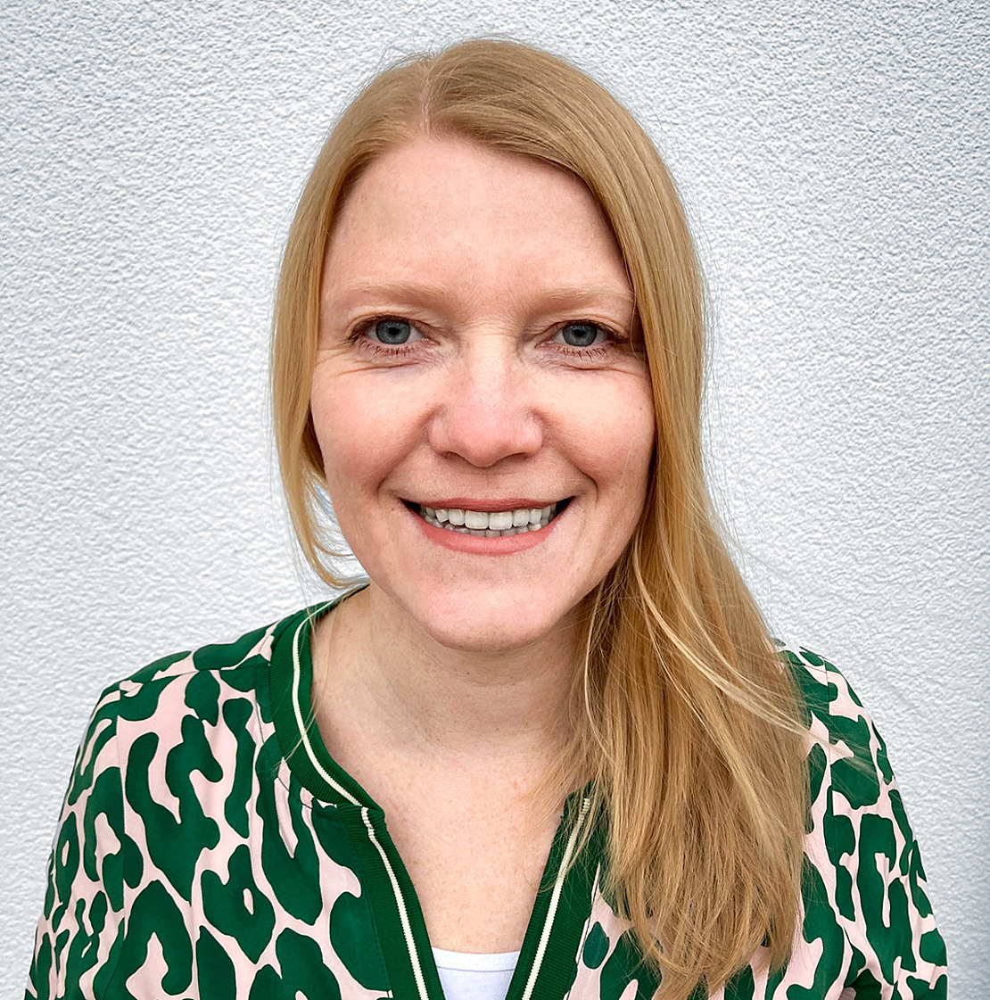 Portrait von Carmen Gorkow Eine Frau mit glattem blondem Haar und blauen Augen lächelt in die Kamera. Sie trägt eine grüne und beigefarbene Bluse mit Tiermuster und steht vor einer weißen, strukturierten Wand.