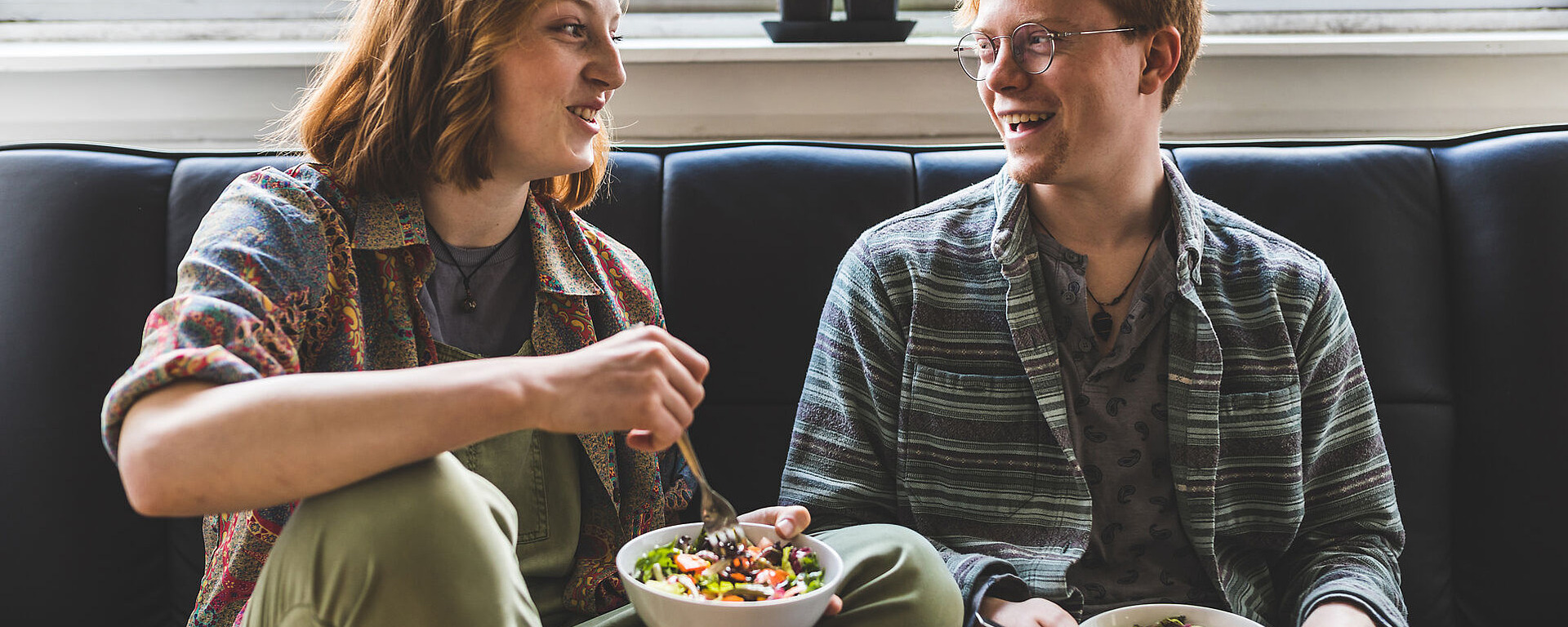 Zwei junge Erwachsene sitzen auf einer Couch am Fenster und lächeln sich an, während sie Schüsseln mit Salat halten und essen. Auf der Fensterbank hinter ihnen ist eine kleine Grünpflanze zu sehen.