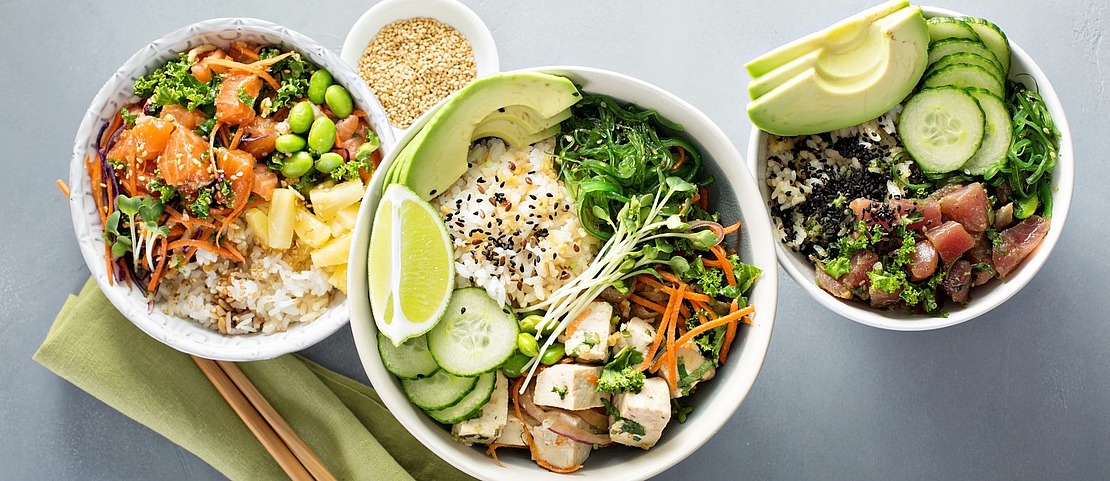Drei Schalen mit Reis, frischem Gemüse, Seetang, Avocado, Gurkenscheiben, Edamame und Fisch oder Tofu stehen auf einer grauen Fläche, daneben Stäbchen und eine kleine Schale mit Sesamkörnern.