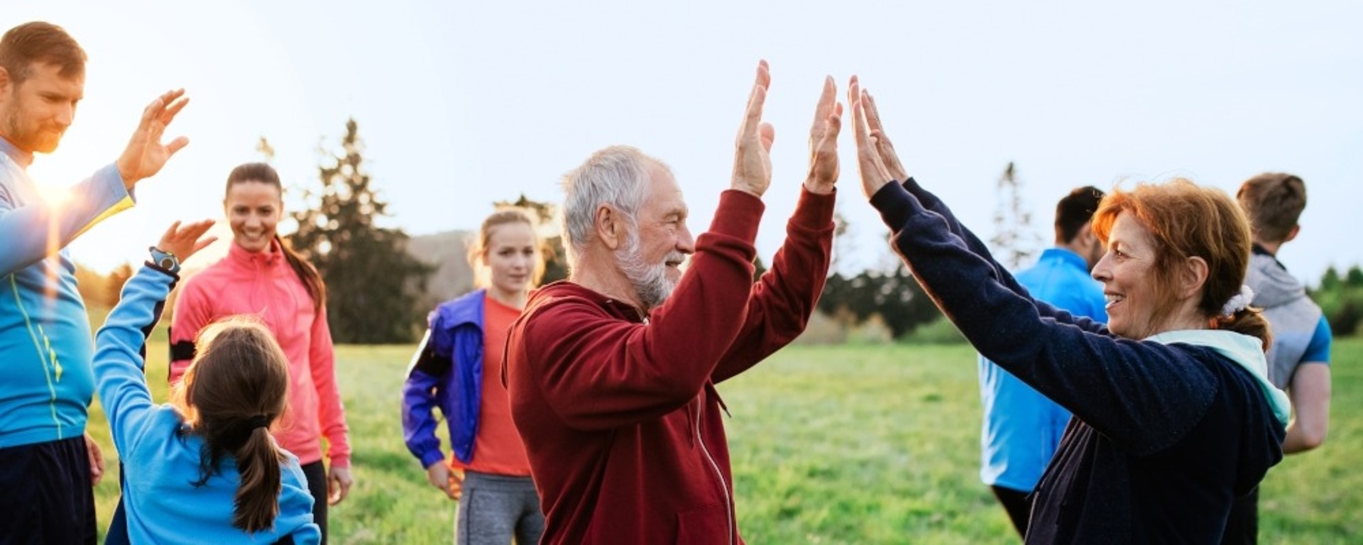 Eine Gruppe von Menschen, darunter Kinder und ältere Erwachsene, steht auf einer Wiese im Freien. Zwei ältere Erwachsene im Vordergrund lächeln und klatschen sich gegenseitig ab, während andere im Hintergrund fröhlich miteinander spielen.