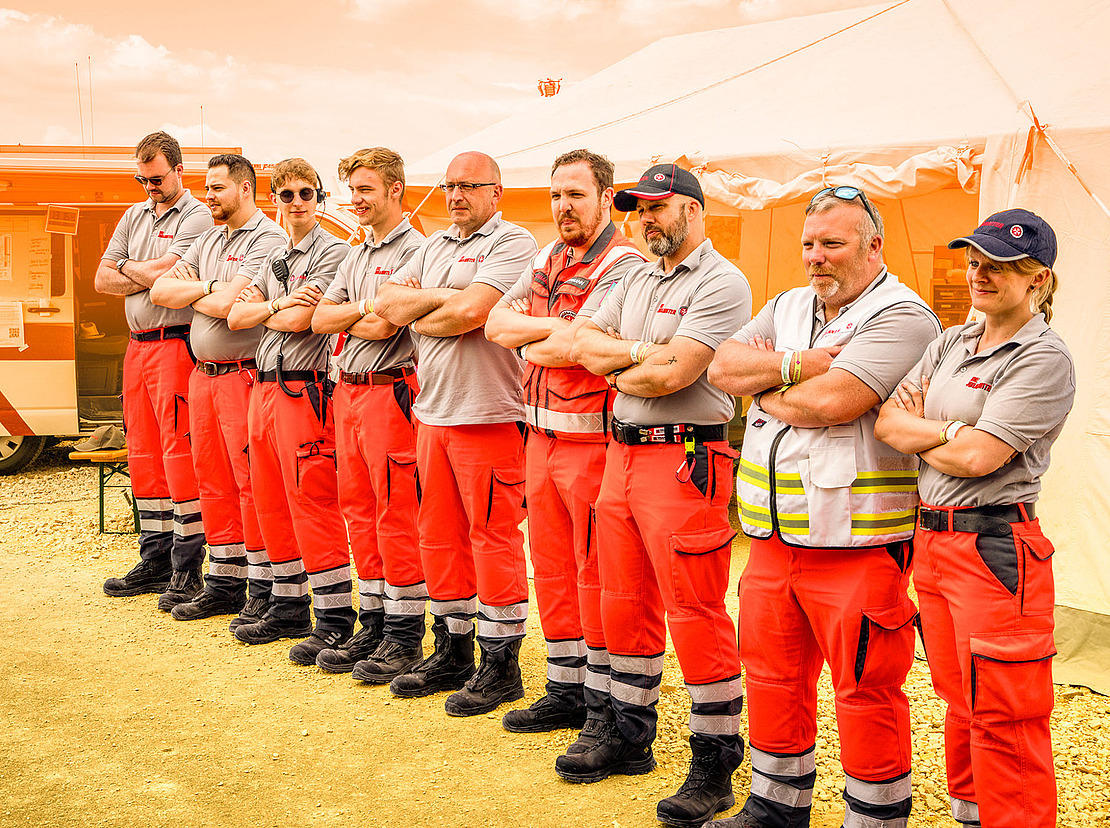 Eine Gruppe von Rettungskräften in passenden grauen Hemden und roten Hosen steht an einem sonnigen Tag mit verschränkten Armen im Freien in der Nähe eines weißen Zeltes und eines Rettungswagens in einer Reihe.