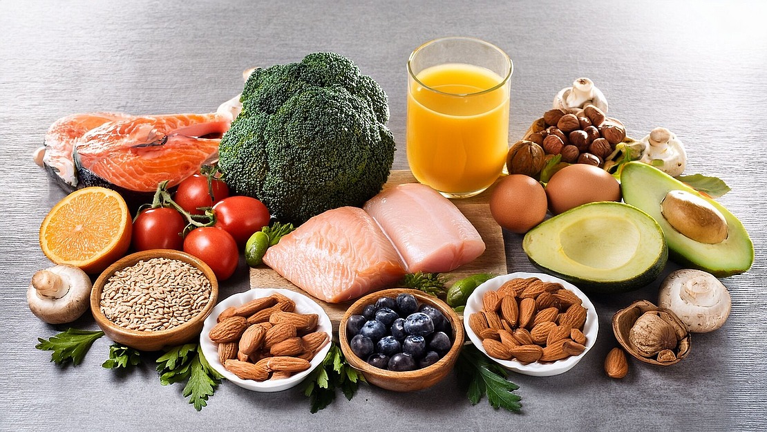 Eine Auswahl gesunder Lebensmittel, darunter Lachs, rohes Hühnchen, Eier, Avocado, Brokkoli, Tomaten, Pilze, Nüsse, Samen, Blaubeeren, eine Orange und ein Glas Orangensaft, auf einem Tisch angerichtet.