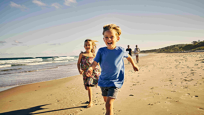 Zwei lächelnde Kinder laufen an einem Sandstrand mit Meereswellen auf der linken Seite auf die Kamera zu. Zwei Erwachsene laufen in der Ferne hinter ihnen, unter einem klaren blauen Himmel.