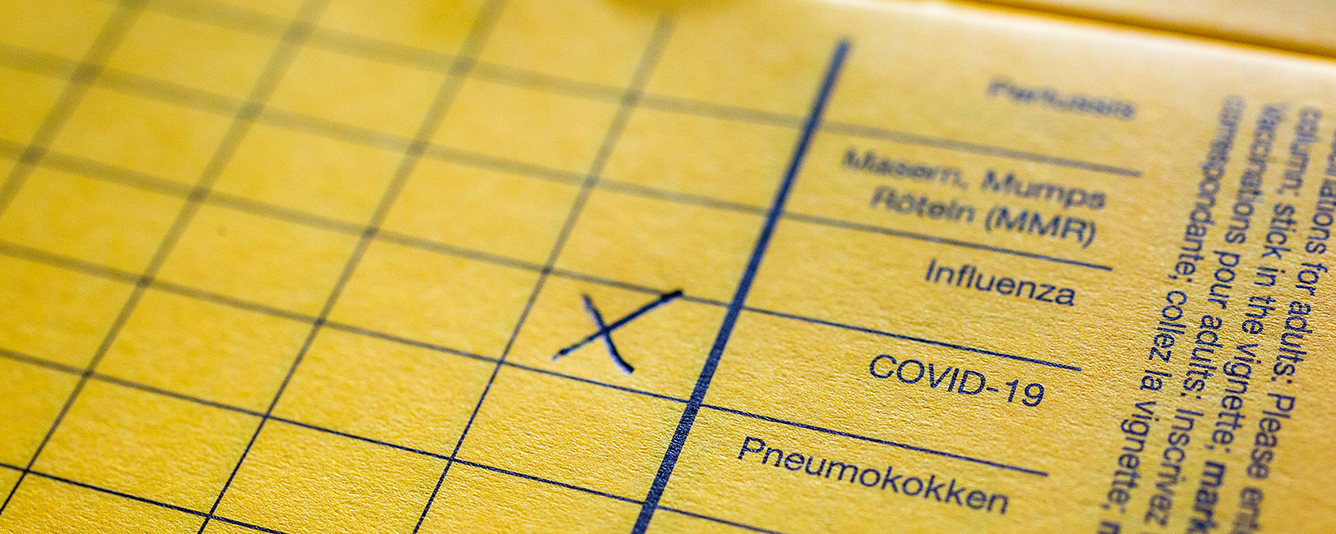 Nahaufnahme eines gelben Impfpasses mit Abschnitten für verschiedene Impfstoffe, darunter COVID-19, mit einem blauen "X" im COVID-19-Feld. Der Text ist in Deutsch und Englisch gedruckt.