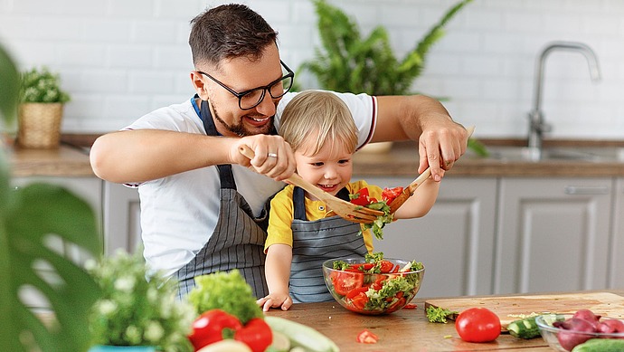 Ein Mann und ein kleines Kind, die beide Schürzen tragen, bereiten gemeinsam einen frischen Salat in einer Küche zu. Sie lächeln und benutzen hölzerne Utensilien, um Gemüse in einer Glasschüssel auf einer Holztheke zu mischen.