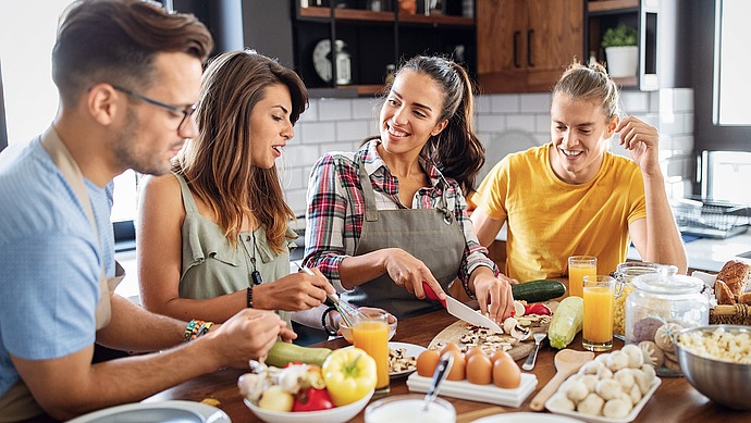 Vier junge Erwachsene sitzen zusammen in einer Küche, bereiten Gemüse und andere Zutaten auf einem Holztisch zu, lächeln und genießen die Gesellschaft des anderen.