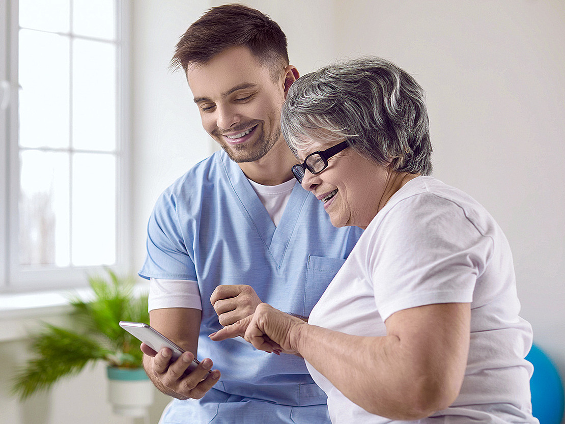 Ein Arzthelfer und eine ältere Patientin schauen gemeinsam auf ein Mobiltelefon. Im Text geht es um Sturzprävention mithilfe von Apps.