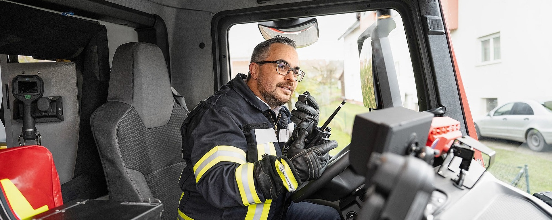 Ein Feuerwehrmann in Uniform und mit Brille sitzt auf dem Fahrersitz eines Feuerwehrautos und spricht in ein Handfunkgerät, während er von Ausrüstung umgeben ist.
