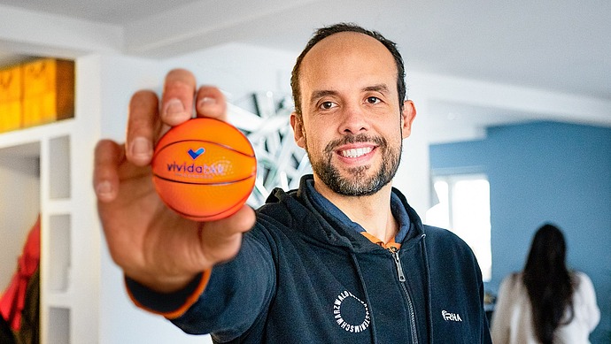 Ein lächelnder Mann in einem dunklen Kapuzenpulli hält einen kleinen orangefarbenen Basketball in die Kamera, während er in einem hell erleuchteten Raum mit einer anderen Person im Hintergrund steht.