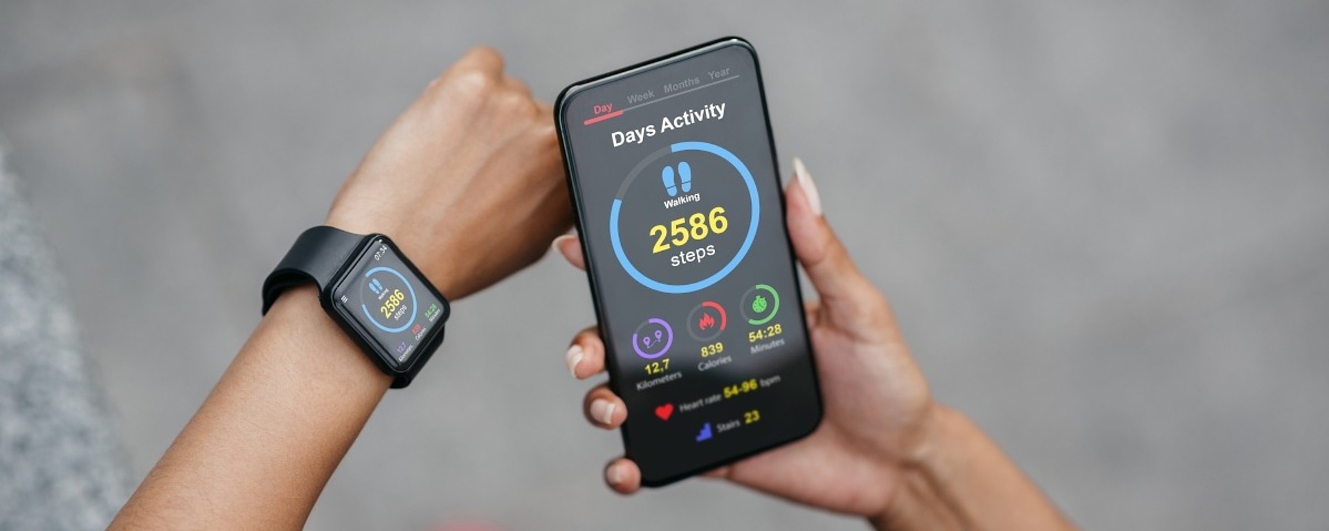 Eine Person schaut auf ein Smartphone, auf dem eine Aktivitätstracker-App mit 2.586 gezählten Schritten angezeigt wird. Sie trägt außerdem eine Smartwatch, die ähnliche Gesundheitsstatistiken anzeigt. Die Person trägt ein schwarzes Outfit und rosa Schuhe und steht auf einer grauen Oberfläche.