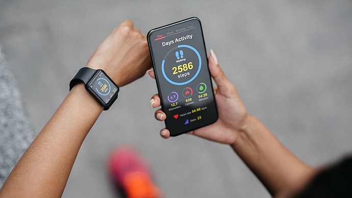 Eine Person schaut auf ein Smartphone, auf dem eine Aktivitätstracker-App mit 2.586 gezählten Schritten angezeigt wird. Sie trägt außerdem eine Smartwatch, die ähnliche Gesundheitsstatistiken anzeigt. Die Person trägt ein schwarzes Outfit und rosa Schuhe und steht auf einer grauen Oberfläche.