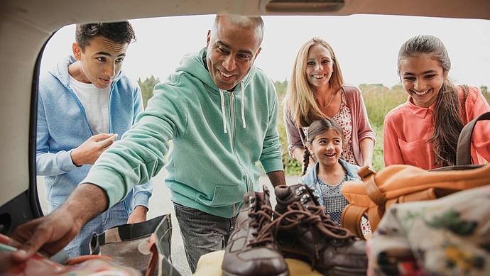 Eine fünfköpfige Familie, darunter zwei Erwachsene und drei Kinder, die lächelnd Gegenstände wie Stiefel und Taschen aus dem Kofferraum eines Autos auslädt, um sich auf eine Aktivität oder ein Abenteuer im Freien vorzubereiten.
