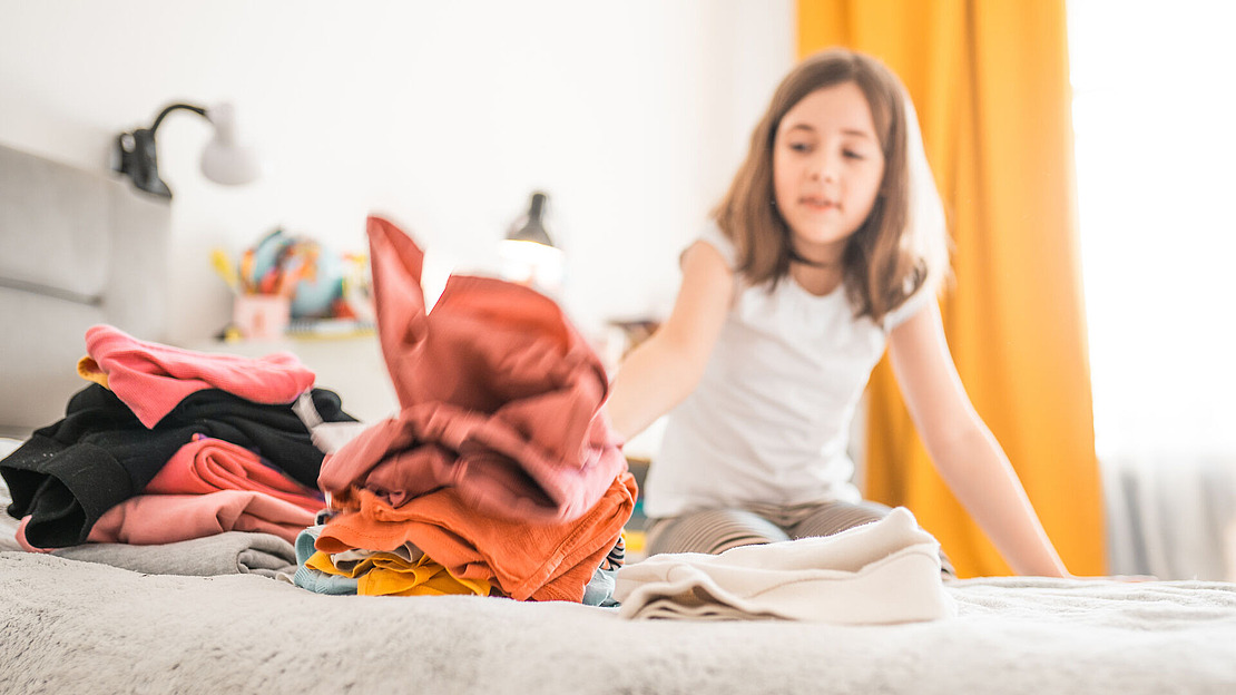 Ein junges Mädchen sitzt auf einem Bett und faltet bunte Kleidung. Im Hintergrund ist ein aufgeräumtes Schlafzimmer mit einer Lampe und gelben Vorhängen zu sehen. Der Fokus liegt auf der gefalteten Kleidung im Vordergrund.