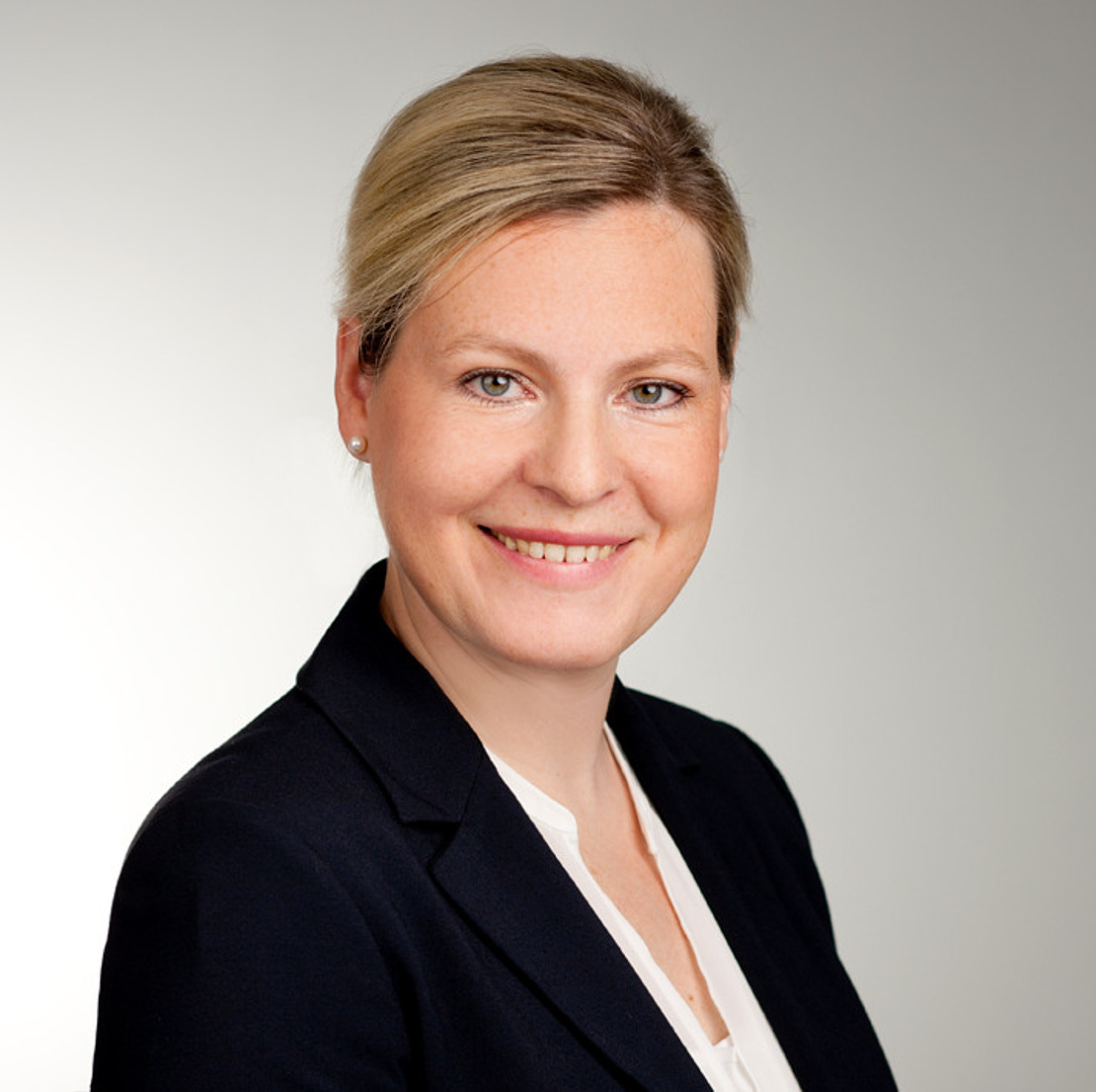 Eine Frau mit blondem Haar in einem Dutt, die einen dunklen Blazer und eine weiße Bluse trägt, lächelt in die Kamera vor einem schlichten hellgrauen Hintergrund.