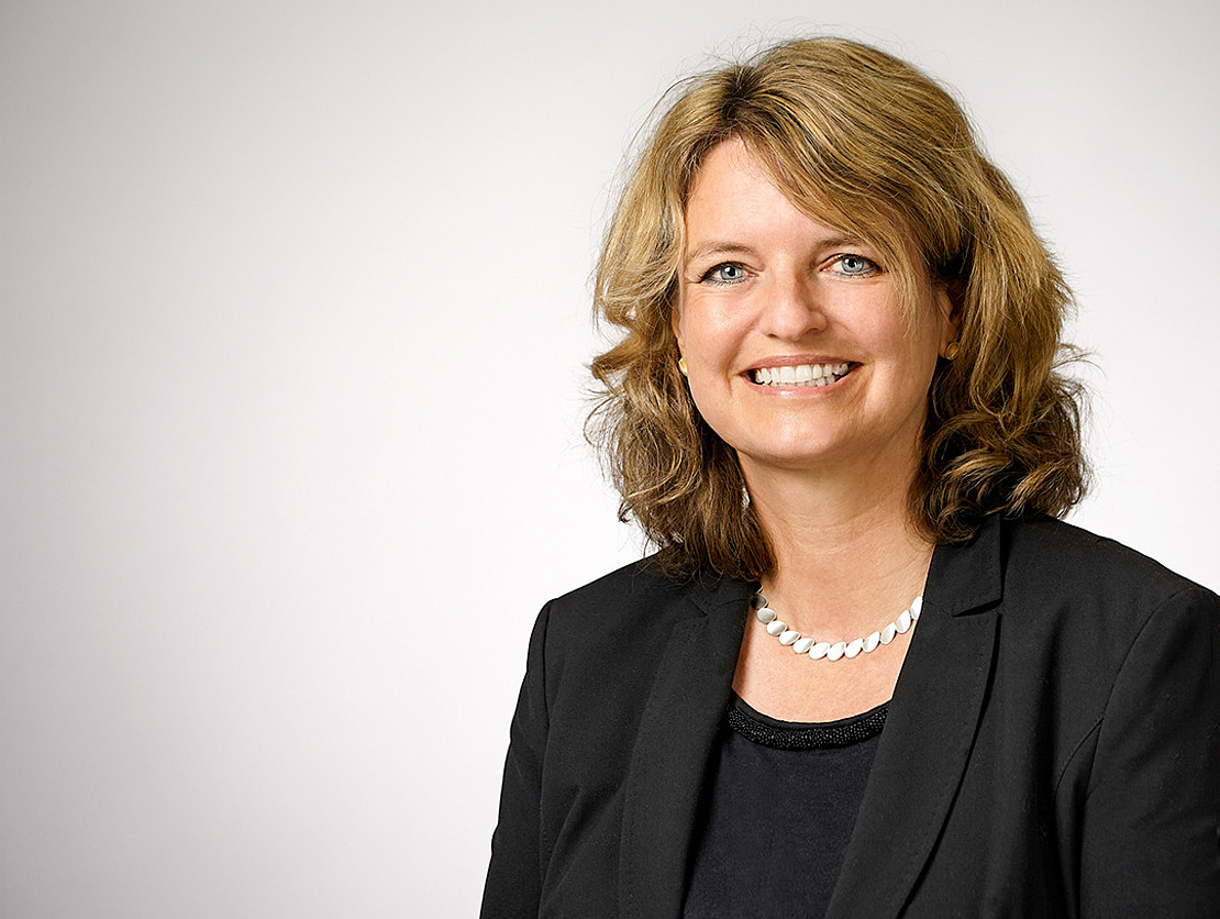 Eine Frau mit gewelltem blondem Haar, die einen schwarzen Blazer, ein schwarzes Oberteil und eine silberne Halskette trägt, lächelt vor einem schlichten hellgrauen Hintergrund in die Kamera.