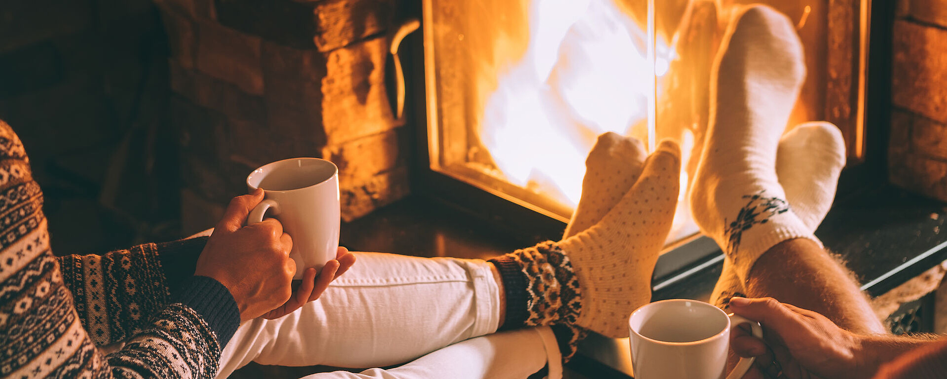 Zwei Personen in kuscheligen Socken sitzen an einem beleuchteten Kamin, jede hält einen weißen Becher. Ihre Beine sind zum Feuer hin ausgestreckt, was eine warme und entspannte Atmosphäre schafft. Pullover und sanfte Beleuchtung tragen zu der gemütlichen Atmosphäre bei.