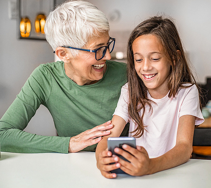 Eine ältere Frau mit kurzen grauen Haaren und Brille lächelt ein junges Mädchen mit langen braunen Haaren an, das an einem Küchentisch ein Smartphone in der Hand hält. Sie sehen beide glücklich und beschäftigt aus.
