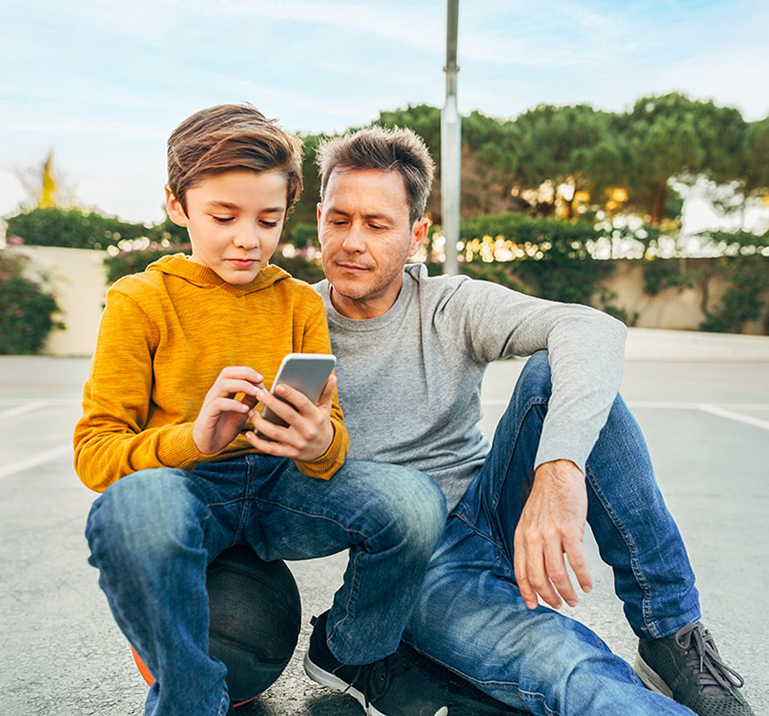 Ein Mann sitzt mit einem Jungen auf dem Asphalt. Der Sohn sitzt auf einem Ball. Der Junge hat ein Smartphone in der Hand, beide schauen in das Smartphone.