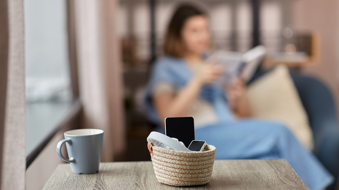 Auf einem Holztisch steht ein Korb mit elektronischen Geräten und Kopfhörern. Daneben steht eine Tasse. Im Hintergrund sitzt eine Person in einem blauen Outfit und liest ein Buch. Der Fokus ist verschwommen.