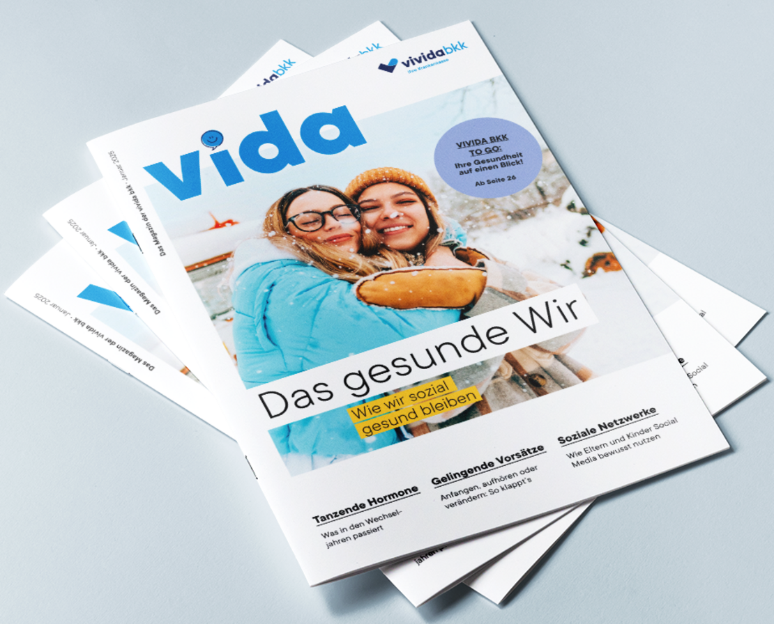 Ein Stapel Zeitschriften mit dem Titel „vida“ und einem Titelbild mit zwei lächelnden Menschen in Winterkleidung. Die Überschrift lautet „Das gesunde Wir“, mit zusätzlichem Text zu Gesundheitsthemen und Features. Der Hintergrund ist eine hellblaue Fläche.